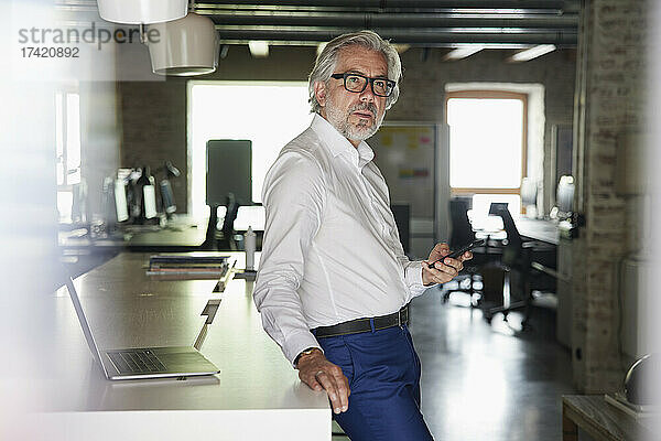 Reifer Geschäftsmann hält Smartphone in der Hand und lehnt am Schreibtisch im Büro