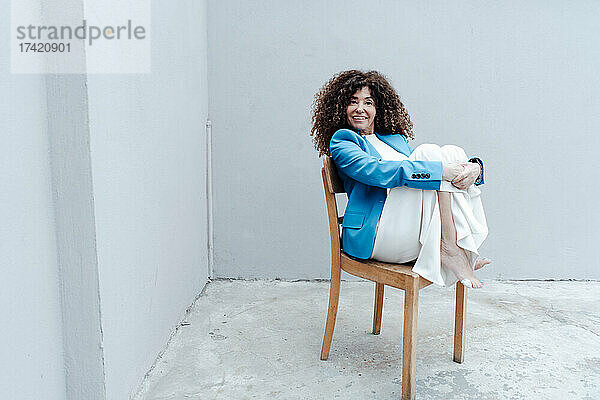 Geschäftsfrau umarmt Knie  während sie auf einem Stuhl vor einer grauen Wand sitzt