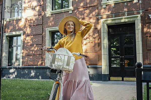 Lächelnde Frau mit Hut steht mit Fahrrad vor Gebäude