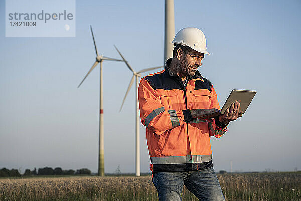 Männlicher Ingenieur nutzt digitales Tablet  während er bei Sonnenuntergang vor Windkraftanlagen steht