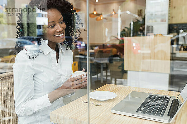 Lächelnde Geschäftsfrau mit Kaffeetasse und Blick auf den Laptop  während sie im Café sitzt