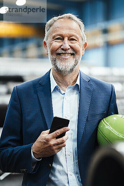 Glücklicher männlicher Geschäftsführer mit Helm und Smartphone in der Industrie