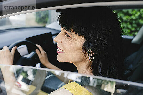 Lächelnde reife Frau schaut beim Fahren eines Elektroautos vom Fenster weg