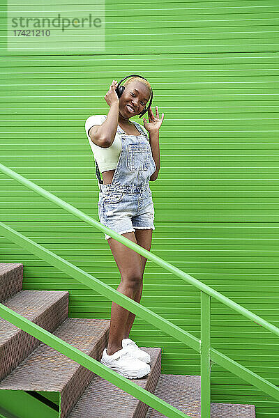 Junge Frau hört Musik über kabellose Kopfhörer auf Stufen vor einer grünen Wand