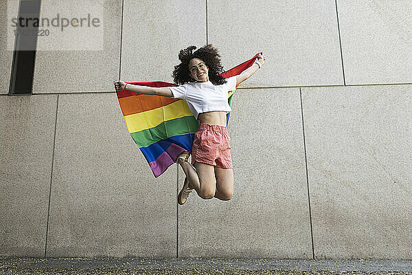 Fröhliche Frau springt  während sie die Regenbogenfahne vor der Wand hält