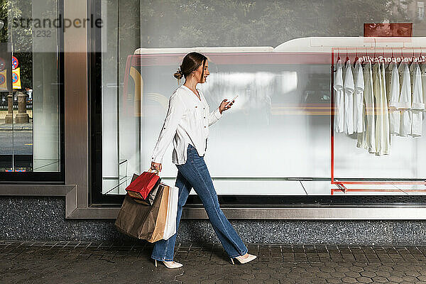 Junge Frau benutzt Smartphone  während sie mit Einkaufstüten durch den Laden geht