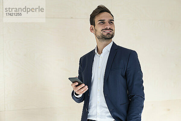 Lächelnder junger Berufstätiger hält sein Mobiltelefon in der Hand  während er vor der Wand steht