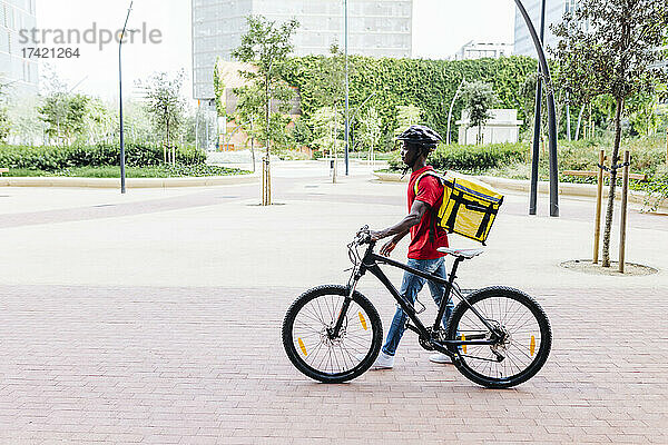 Lieferbote trägt Helm  während er Fahrrad auf Fußweg schiebt