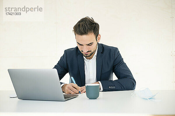 Junger Geschäftsmann schreibt  während er mit Laptop am Schreibtisch im Büro sitzt