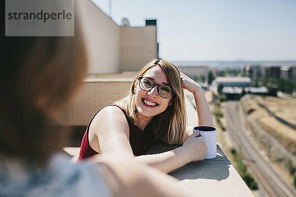 Lächelnde Geschäftsfrau schaut Kollegen an  während sie sich an einem sonnigen Tag an eine Stützmauer lehnt