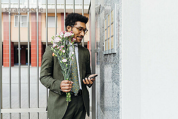Geschäftsmann hält Blumenstrauß und Mobiltelefon in der Hand  während er an der Wand steht