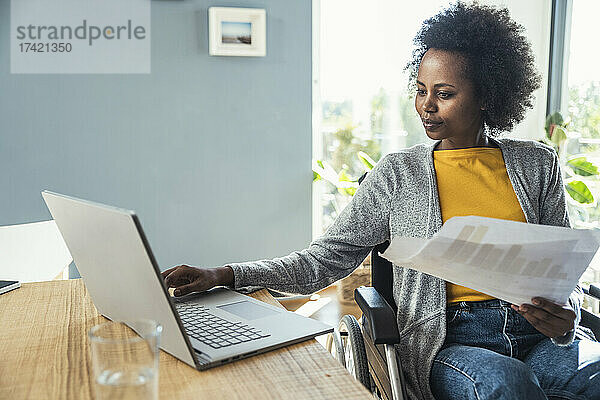 Behinderte Geschäftsfrau hält Dokument in der Hand  während sie im Heimbüro am Laptop arbeitet