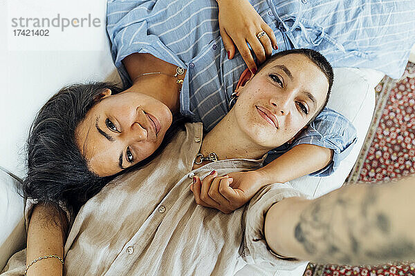 Smiling lesbian couple taking selfie while lying on sofa at home