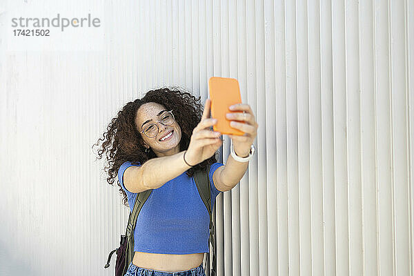 Lächelnde Frau macht Selfie mit Smartphone an weißer Wand