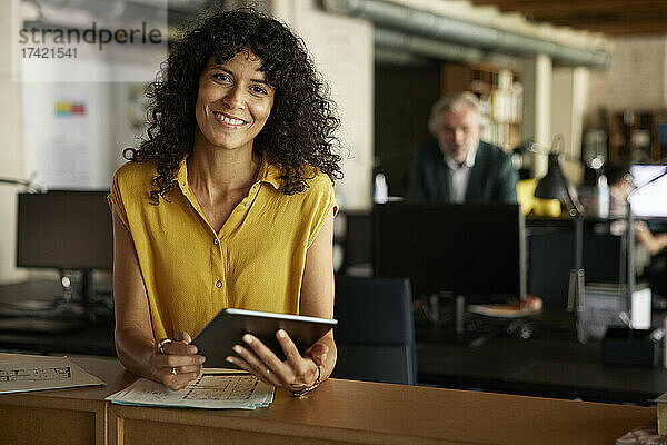 Lächelnde Geschäftsfrau mit digitalem Tablet lehnt am Tisch im Büro