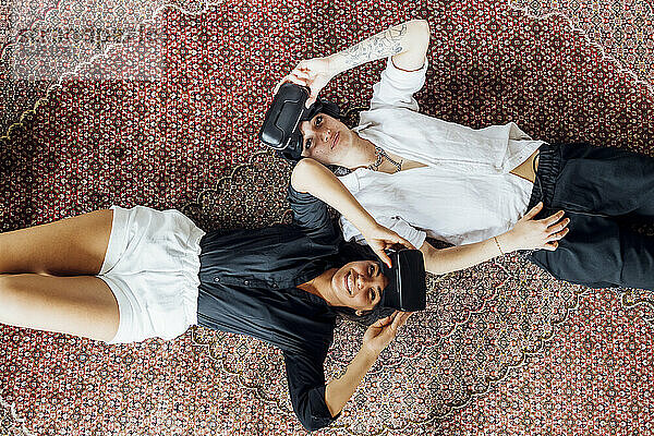 Smiling lesbian couple removing virtual reality simulators while lying on carpet at home