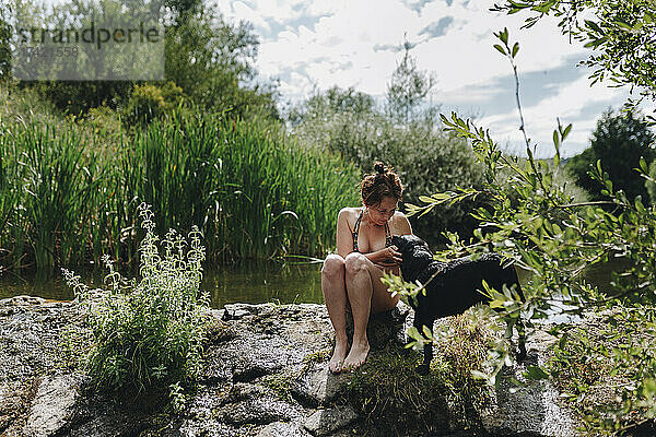 Tierbesitzerin streichelt Hund  während sie auf einem Felsen an einem Teich sitzt