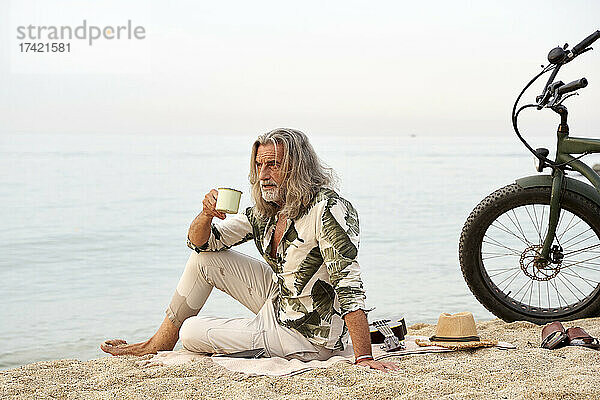 Mann mit langen grauen Haaren hält einen Becher in der Hand  während er mit dem Fahrrad am Strand sitzt