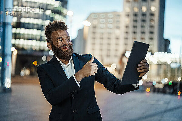 Lächelnder männlicher Freiberufler gestikuliert mit dem Daumen nach oben  während er ein Selfie mit einem digitalen Tablet macht