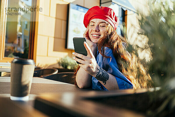 Lächelnde junge Frau benutzt Smartphone im Straßencafé
