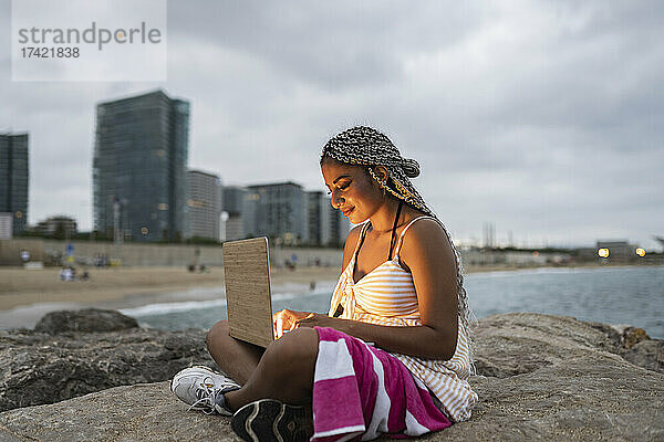 Junge Frau benutzt Laptop  während sie bei Sonnenuntergang auf einem Felsen am Strand sitzt