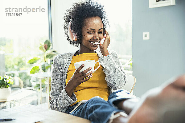 Glückliche Frau hört Musik über Kopfhörer  während sie zu Hause Kaffee trinkt