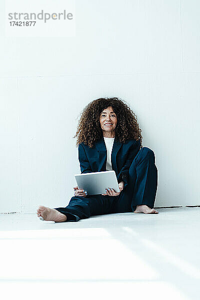 Geschäftsfrau mit digitalem Tablet trägt übergroßen Anzug  während sie vor der Wand sitzt