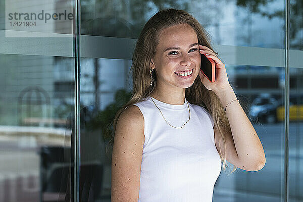 Glückliche junge Freiberuflerin  die vor einer Glaswand mit dem Handy spricht