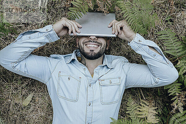 Sorgloser Mann liegt auf Pflanzen und bedeckt Gesicht mit digitalem Tablet im Wald