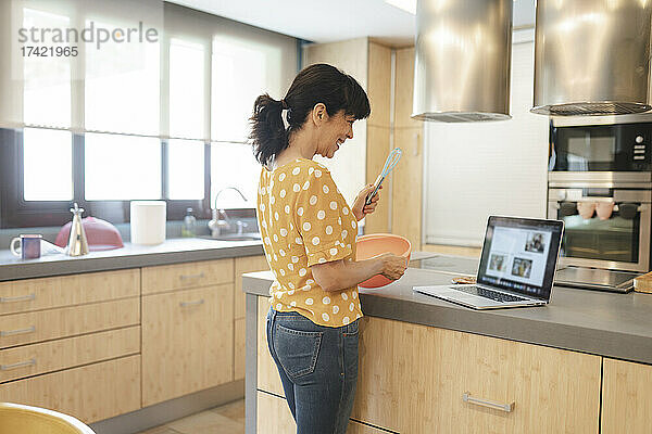 Glückliche Frau bereitet Essen zu  während sie in der heimischen Küche am Laptop lernt