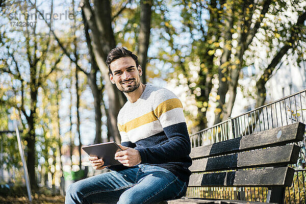Lächelnder junger Mann hält ein digitales Tablet in der Hand  während er an einem sonnigen Tag auf einer Bank im Park sitzt