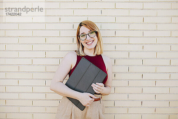 Lächelnde Geschäftsfrau hält digitales Tablet in der Hand  während sie vor einer Ziegelwand steht