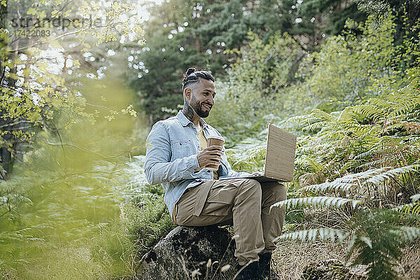 Lächelnder Mann mit wiederverwendbarer Kaffeetasse und Laptop im Wald