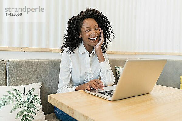 Fröhliche Geschäftsfrau mit Hand am Kinn nimmt über Laptop an Videoanruf im Café teil