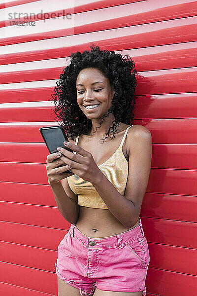 Lächelnde Frau mit lockigem Haar benutzt Smartphone vor rotem Verschluss