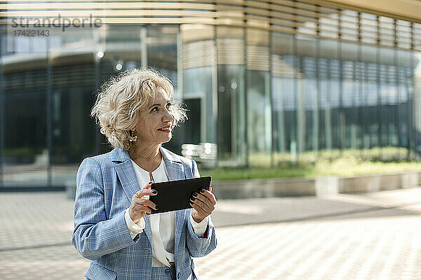 Lächelnde blonde Geschäftsfrau hält digitales Tablet vor Bürogebäude
