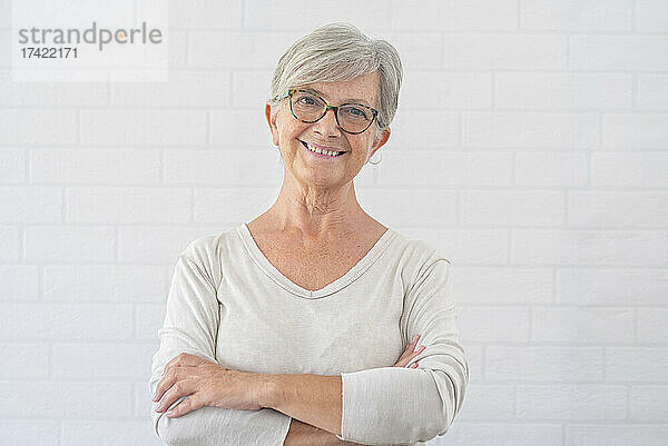 Lächelnde ältere Frau mit verschränkten Armen vor weißer Wand