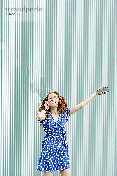 Lächelnde junge rothaarige Frau genießt Musik vor blauer Wand