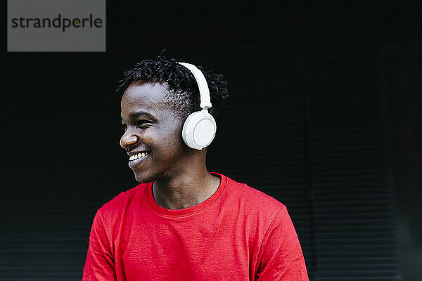 Glücklicher junger Mann  der vor einer schwarzen Wand Musik über Kopfhörer hört