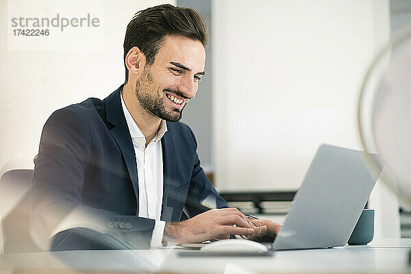Glücklicher junger Geschäftsmann benutzt Laptop  während er am Schreibtisch im Büro sitzt
