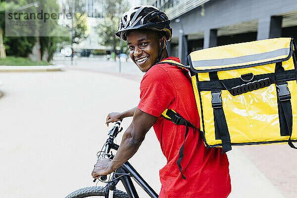 Glücklicher junger Lieferbote mit Rucksack  der auf dem Fahrrad sitzt
