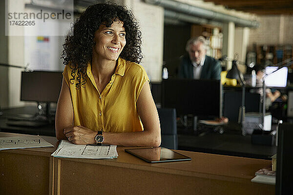 Lächelnde  lockige Geschäftsfrau mit Dokumenten und digitalem Tablet lehnt am Tisch im Büro