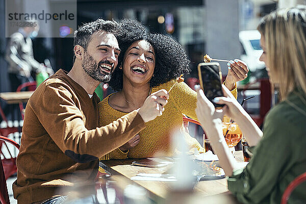 Frau fotografiert fröhliche männliche und weibliche Freunde  die im Restaurant Essen zeigen
