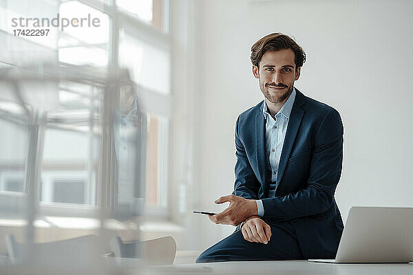 Lächelnder Geschäftsmann mit Mobiltelefon sitzt im Büro
