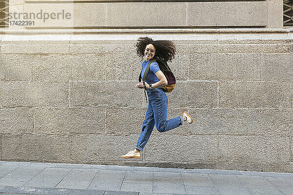 Glückliche Frau mit Rucksack springt auf Fußweg vor Mauer