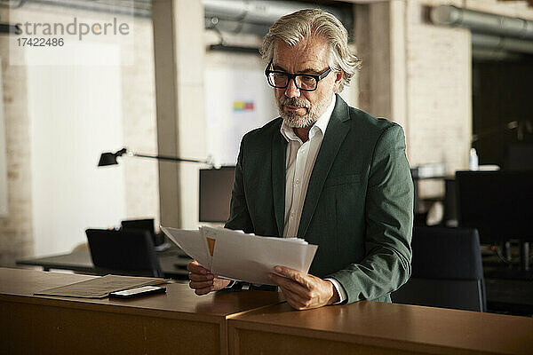 Geschäftsmann mit grauen Haaren analysiert Dokumente  während er am Tisch im Büro steht