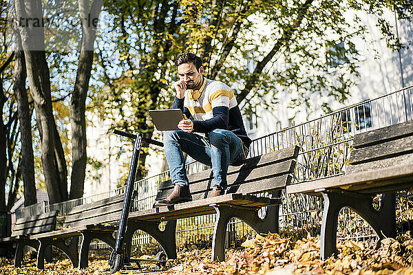 Junger Mann nutzt digitales Tablet  während er an sonnigen Tagen mit einem Elektroroller auf einer Bank im Park sitzt