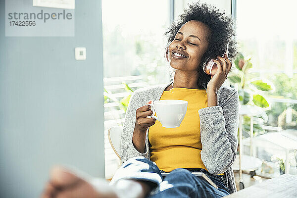 Junge Frau mit geschlossenen Augen hält Kaffeetasse in der Hand  während sie im Wohnzimmer Musik hört