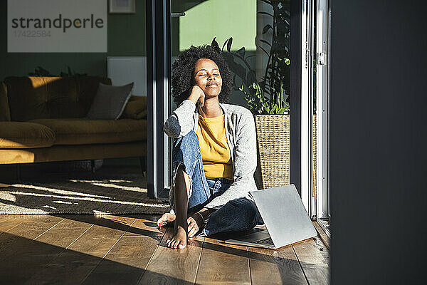 Frau mit geschlossenen Augen entspannt sich im Sonnenlicht  während sie zu Hause auf dem Boden sitzt