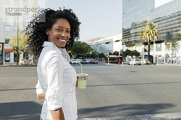 Lächelnde Geschäftsfrau hält Getränk in der Hand  während sie an der Stadtstraße steht